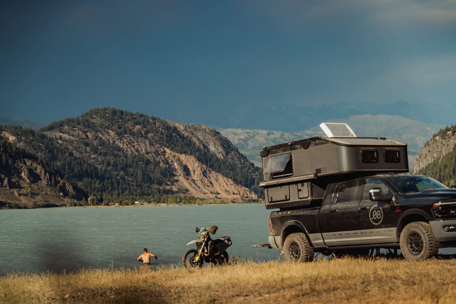 A Limited Edition Scout Camper truck and motorcycle are parked by a lake with mountains in the background. A person is swimming in the water under a cloudy Sitka sky.