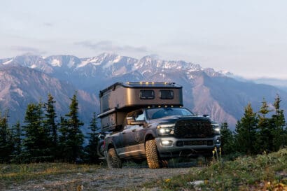 A rugged Scout Camper pickup truck in the Limited Edition Sitka trim is parked on a dirt path surrounded by pine trees, with majestic snow-capped mountains in the background under a clear sky.