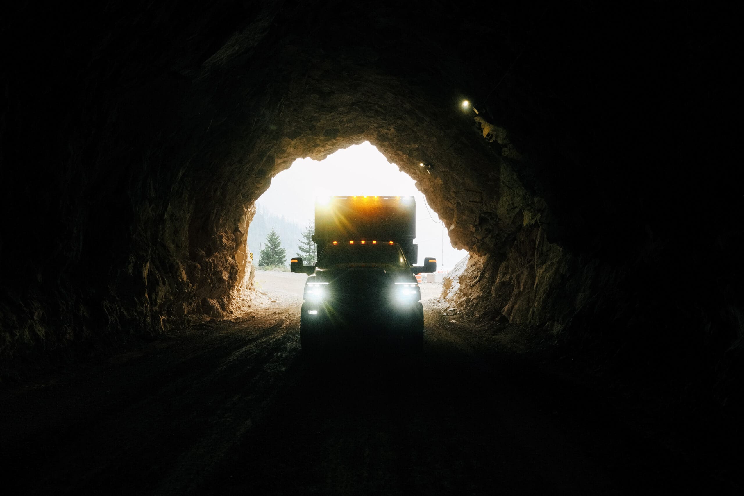 A large truck with headlights on drives out of a dark tunnel toward daylight, framed by rocky tunnel walls and a glimpse of trees outside—a Scout Camper perched on its bed, ready for limitless adventure.