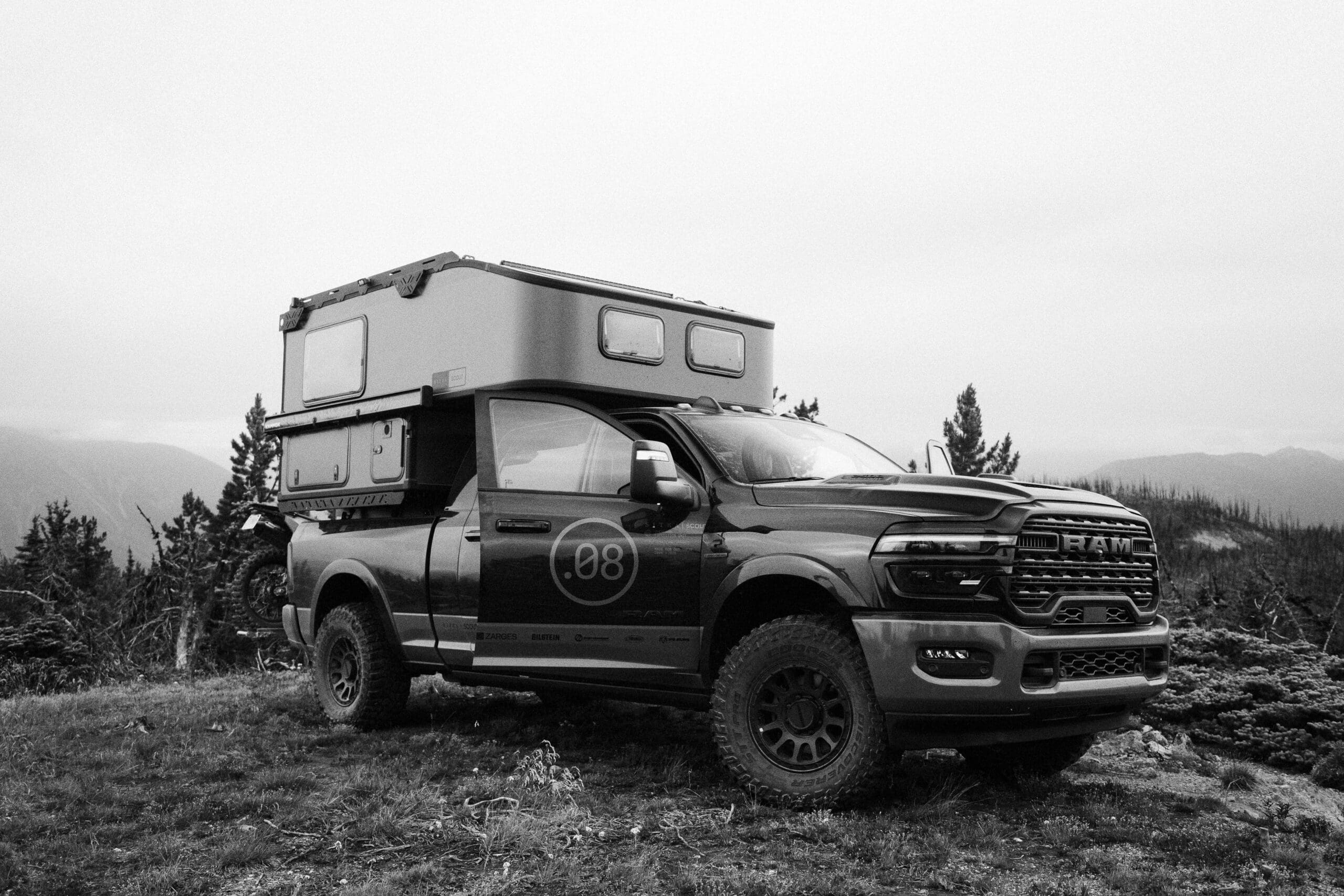 A black and white photo of a pickup truck with large tires and a Limited Edition Scout Camper attached, parked on grass with trees and mountains in the background.