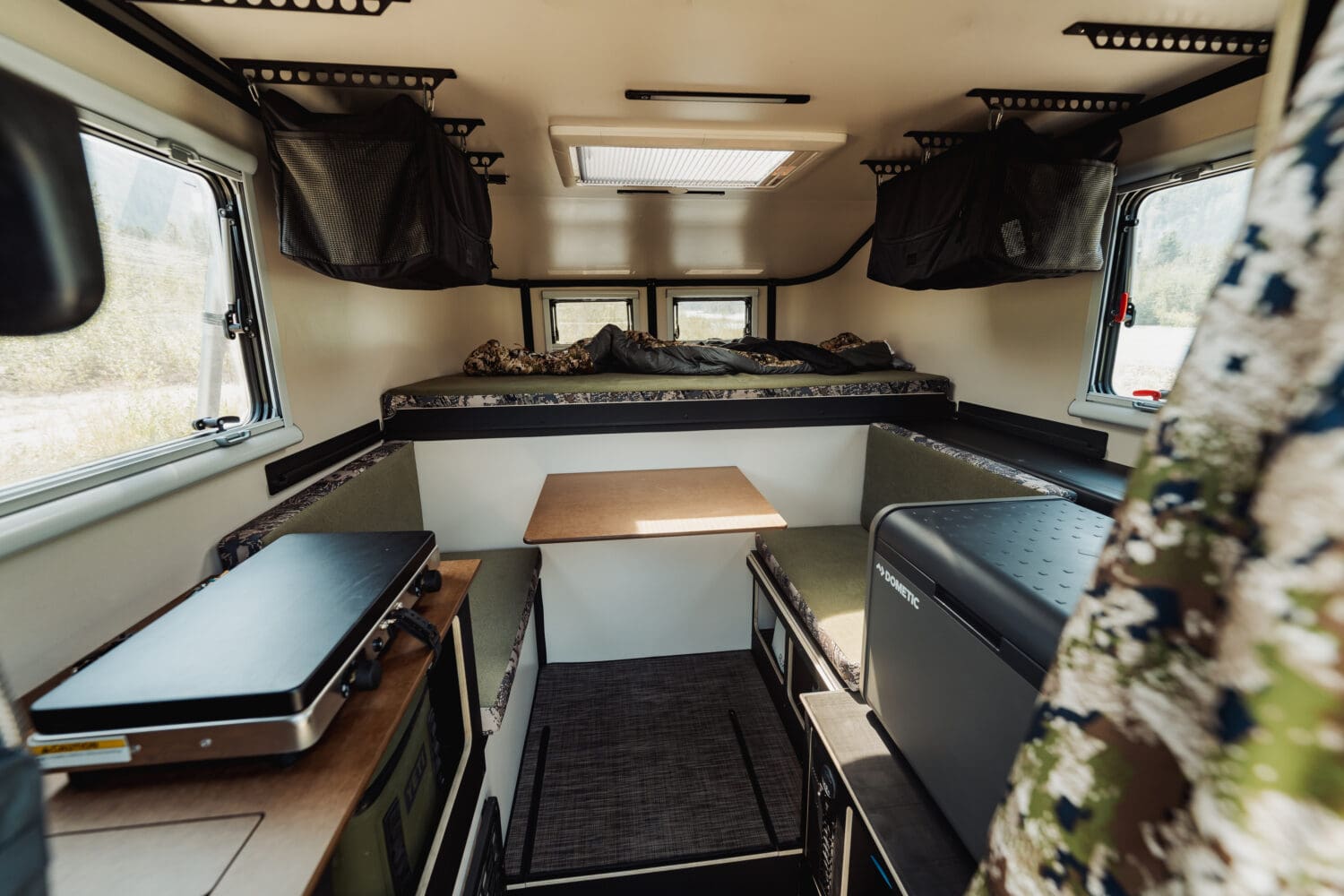 The interior of the Sitka Scout Camper Limited Edition features a small bed in the back, a foldable table, benches, a portable stove, and storage compartments, with windows on both sides letting in natural light.