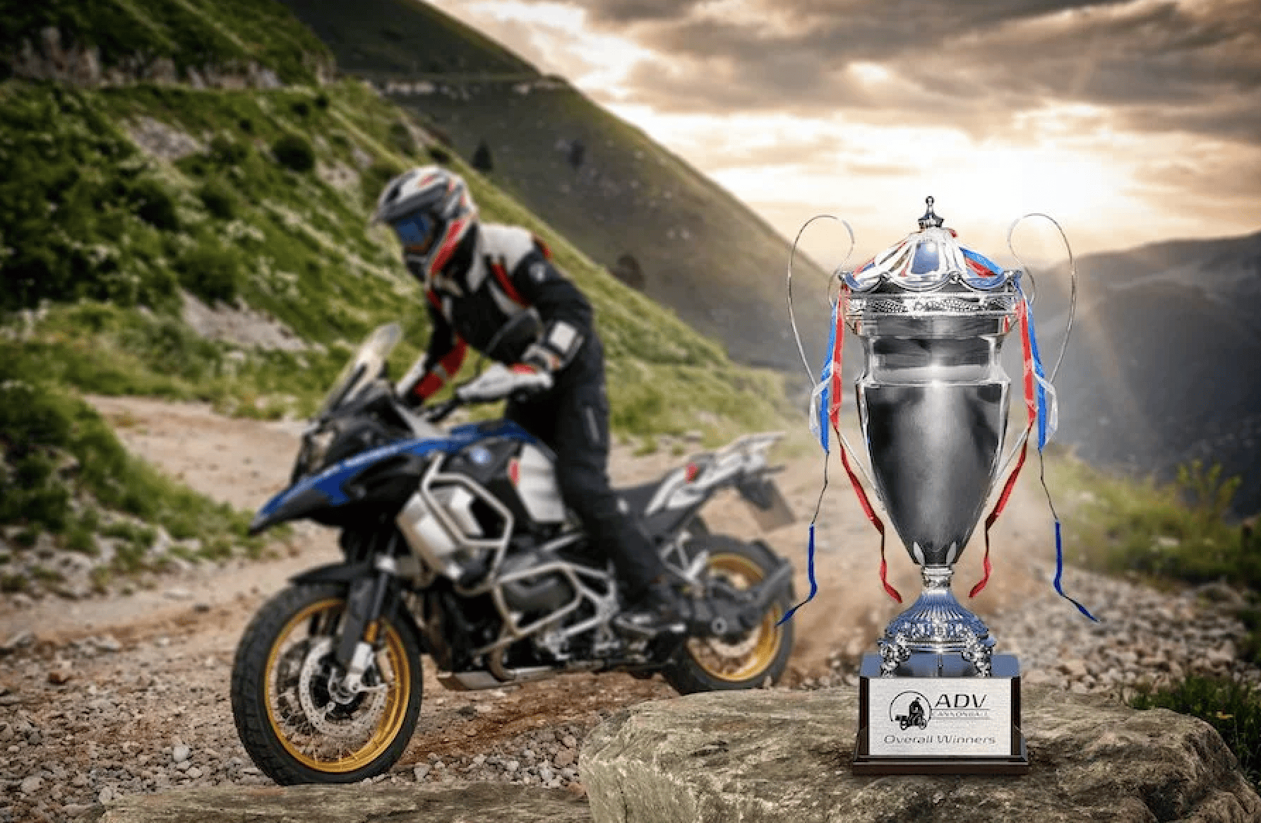 A motorcyclist in riding gear on a dirt path, with a large trophy labeled ADV Overall Winners placed on rocks in the foreground. Set against mountains and a dramatic sky, the scene captures the spirit of Fall at Overland Events, where adventure and achievement come together.