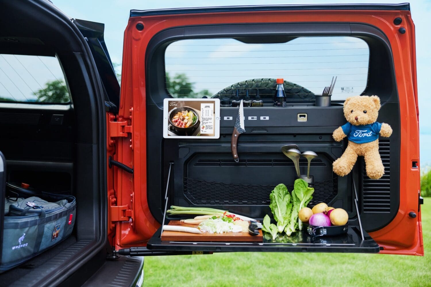 An open orange Ford Bronco EV trunk with a tailgate kitchen setup, ingredients on a cutting board, cooking utensils, a recipe book, condiments, and a Ford teddy bear hanging on the door.
