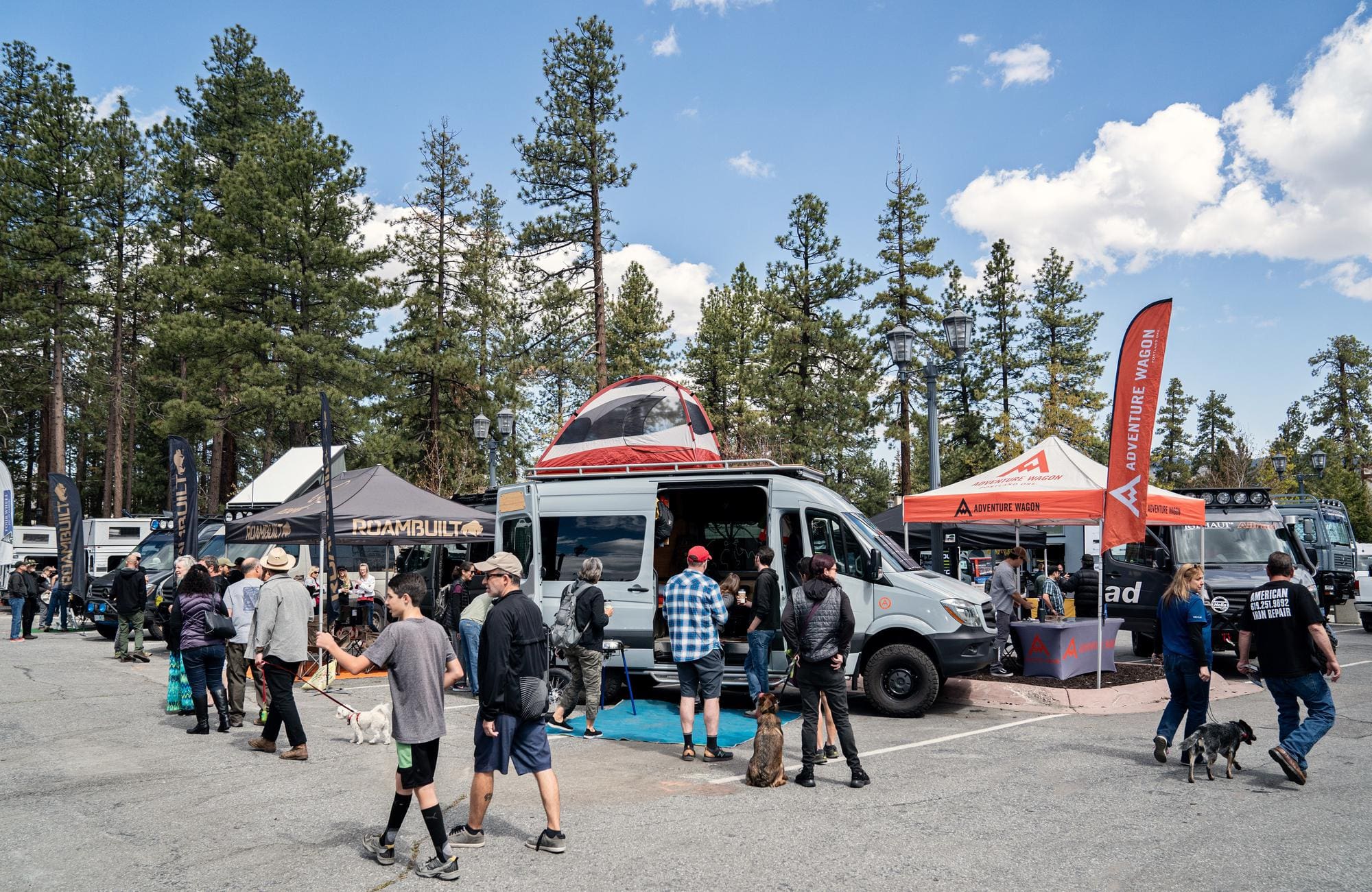 People gather around camper vans and tents at an outdoor expo surrounded by tall pine trees. Some attendees walk dogs, while others explore exhibits and gear displays under banners and flags—a lively scene typical of overland events and rallies.