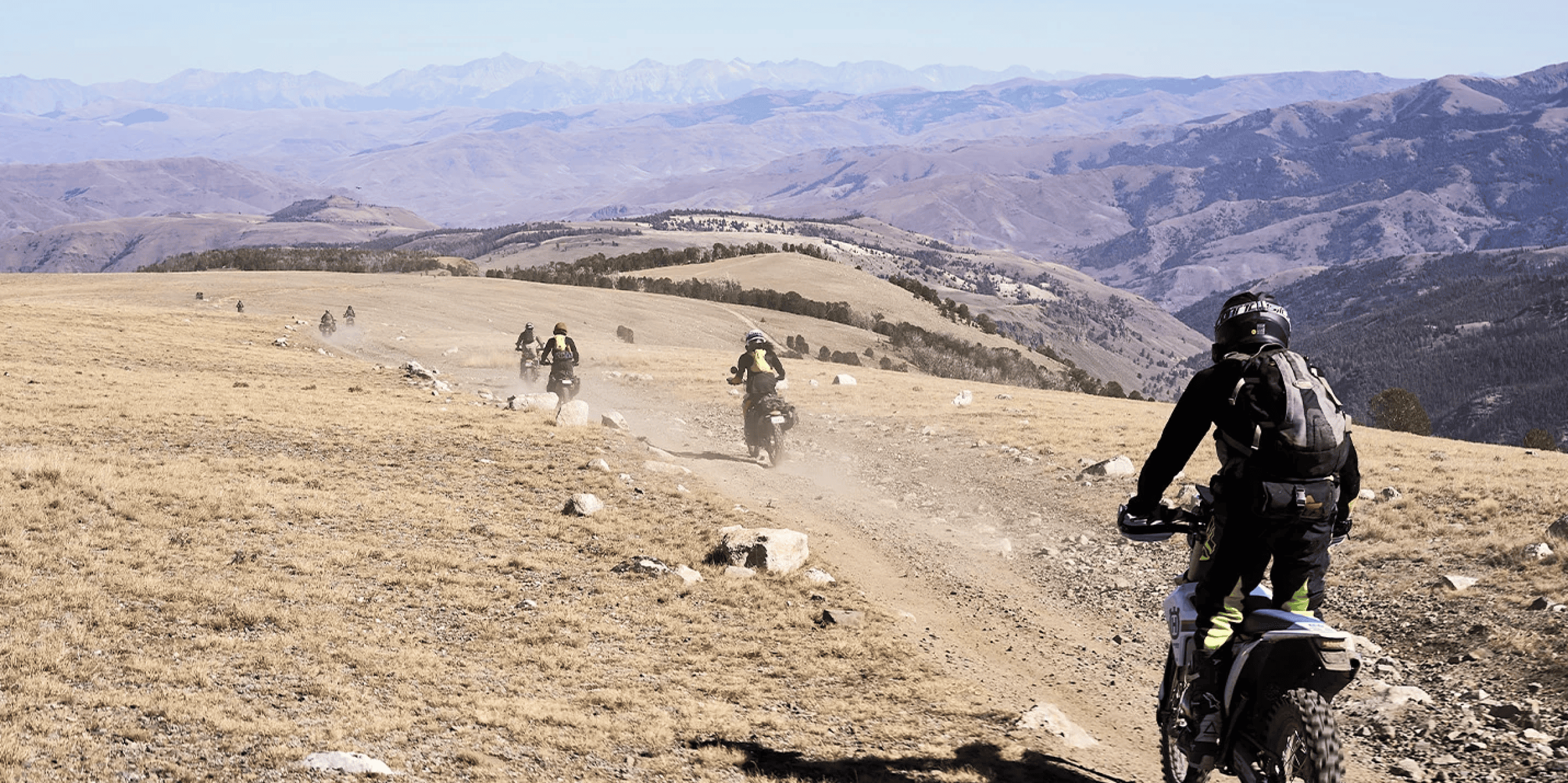 A group of motorcyclists ride on a dusty dirt trail through a mountainous, dry landscape under a clear sky with distant peaks in the background, capturing the adventurous spirit of Overland Rallies and autumn Overland Events.