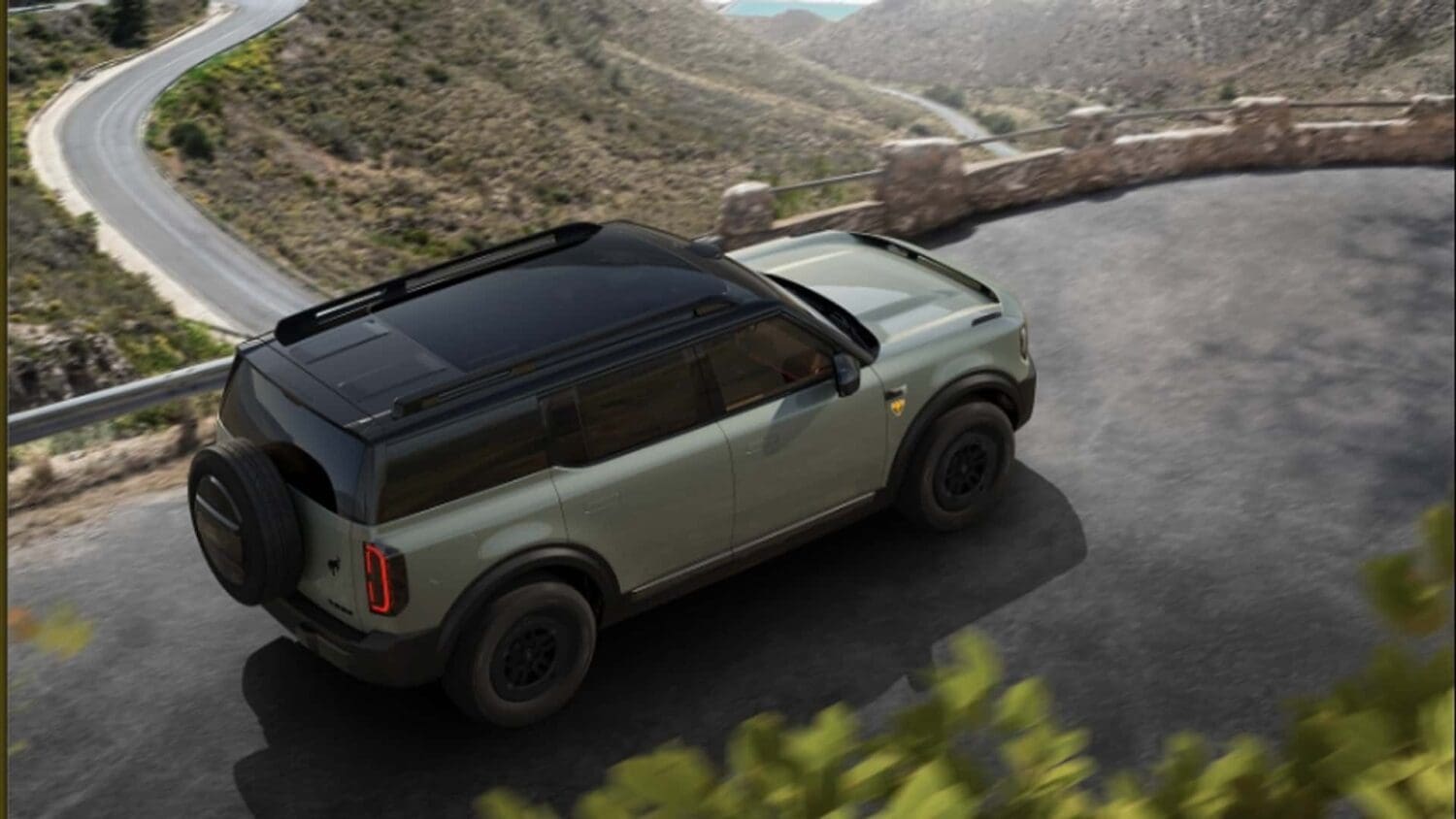 A green Ford Bronco EV with a black roof drives along a winding mountain road with scenic views, surrounded by rocky hills and some greenery.