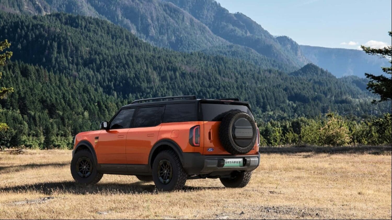 An orange Ford Bronco EV SUV with a spare tire on the back is parked on dry grass with mountains and pine forests in the background under a clear sky.