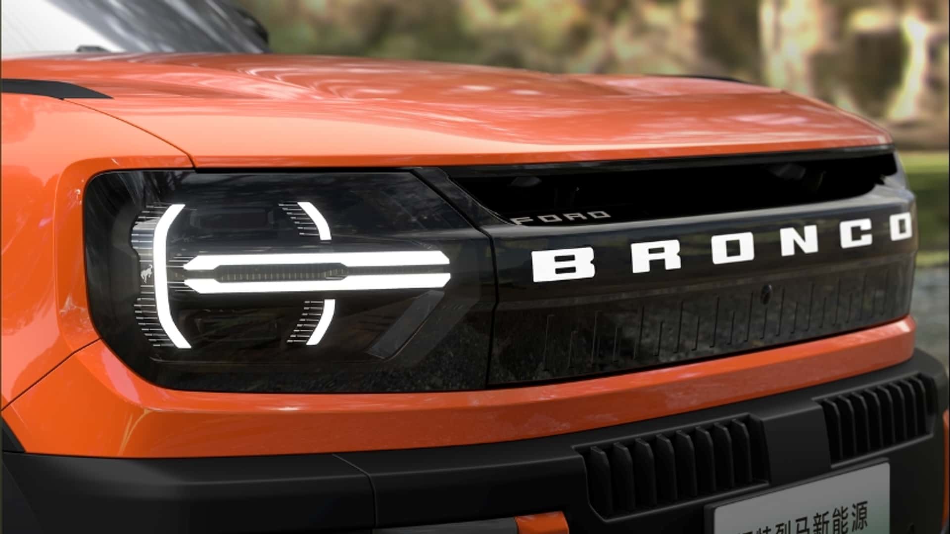 Close-up of an orange Ford Bronco EV’s front, showcasing the bold BRONCO lettering on the black grille and modern LED headlights, with a blurred outdoor background—highlighting the striking presence of this electric SUV.