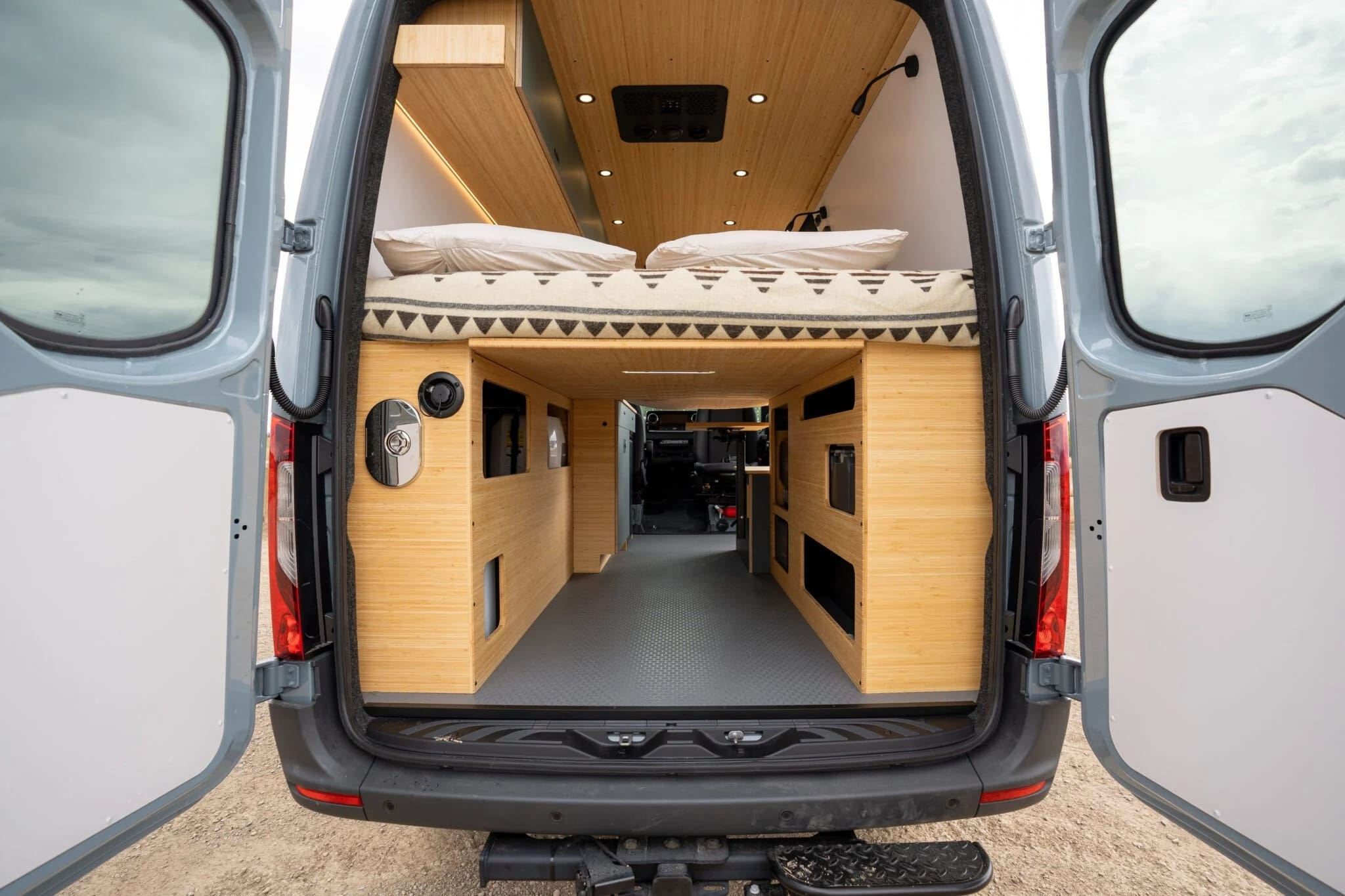 The image shows the interior of a Geotrek Flatiron camper van with wooden furnishings, a bed above storage cabinets, and an open back door. The space is neatly organized, featuring overhead lighting and a minimalist design.