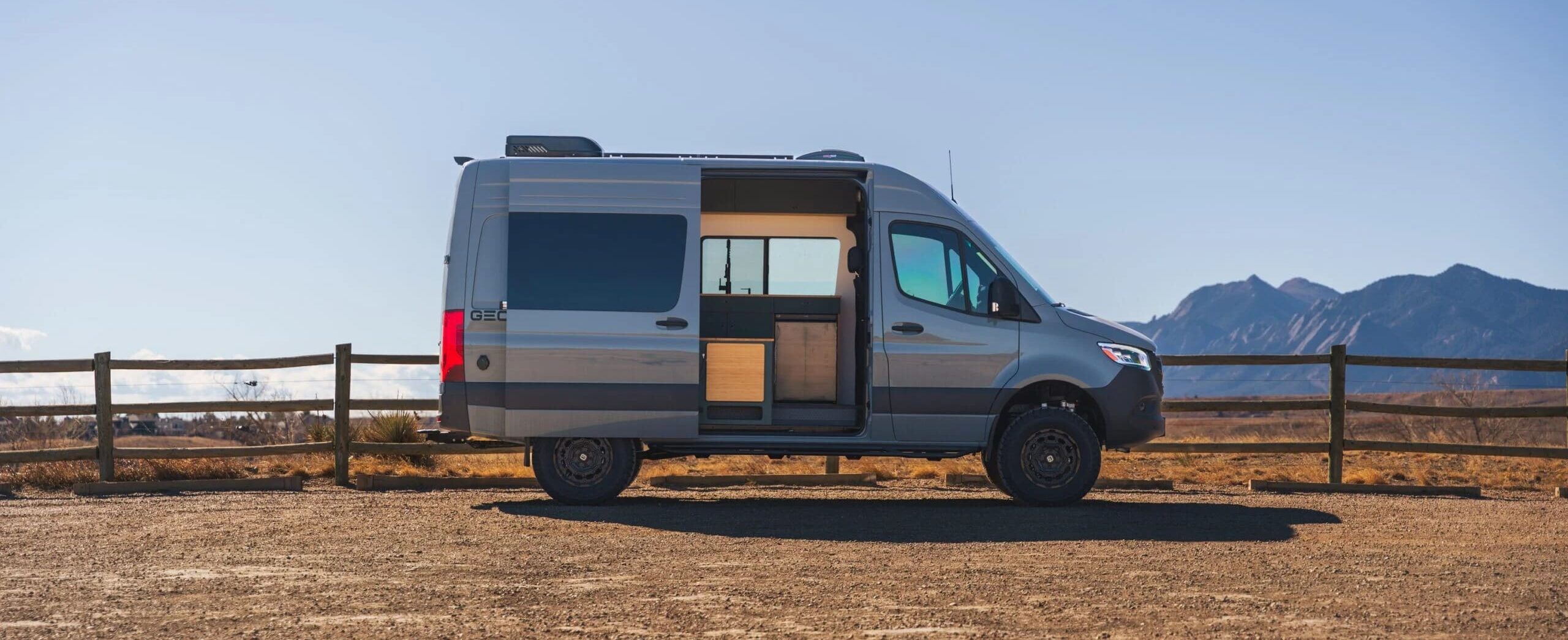 A silver Geotrek Flatiron camper van with its side door open is parked on a dirt lot near a wooden fence, with mountains and a clear blue sky in the background.