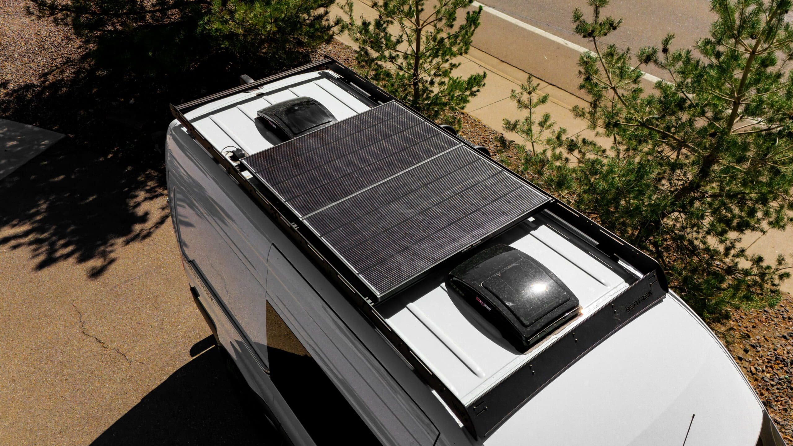 A white Geotrek Flatiron van with two rooftop vents and large black solar panels is parked near trees on a paved surface.