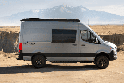 A gray Geotrek Flatiron camper van labeled GEOTREK is parked on a dirt road in a desert landscape, with mountains visible in the background under a clear sky.