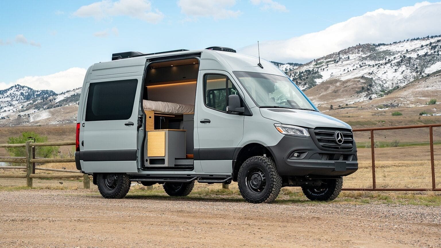 A gray Mercedes-Benz Sprinter Geotrek Flatiron camper van with its side door open, revealing a cozy interior. The van is parked on a dirt area, with mountains and a wooden fence in the background under a partly cloudy sky.