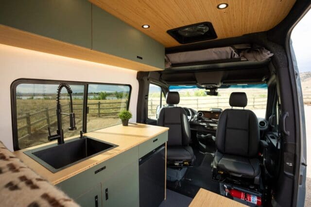 Modern Geotrek Flatiron camper van interior with green cabinets, a black sink with a tall faucet, wood countertops, two front seats, and a large window showing a fence and outdoor scenery.