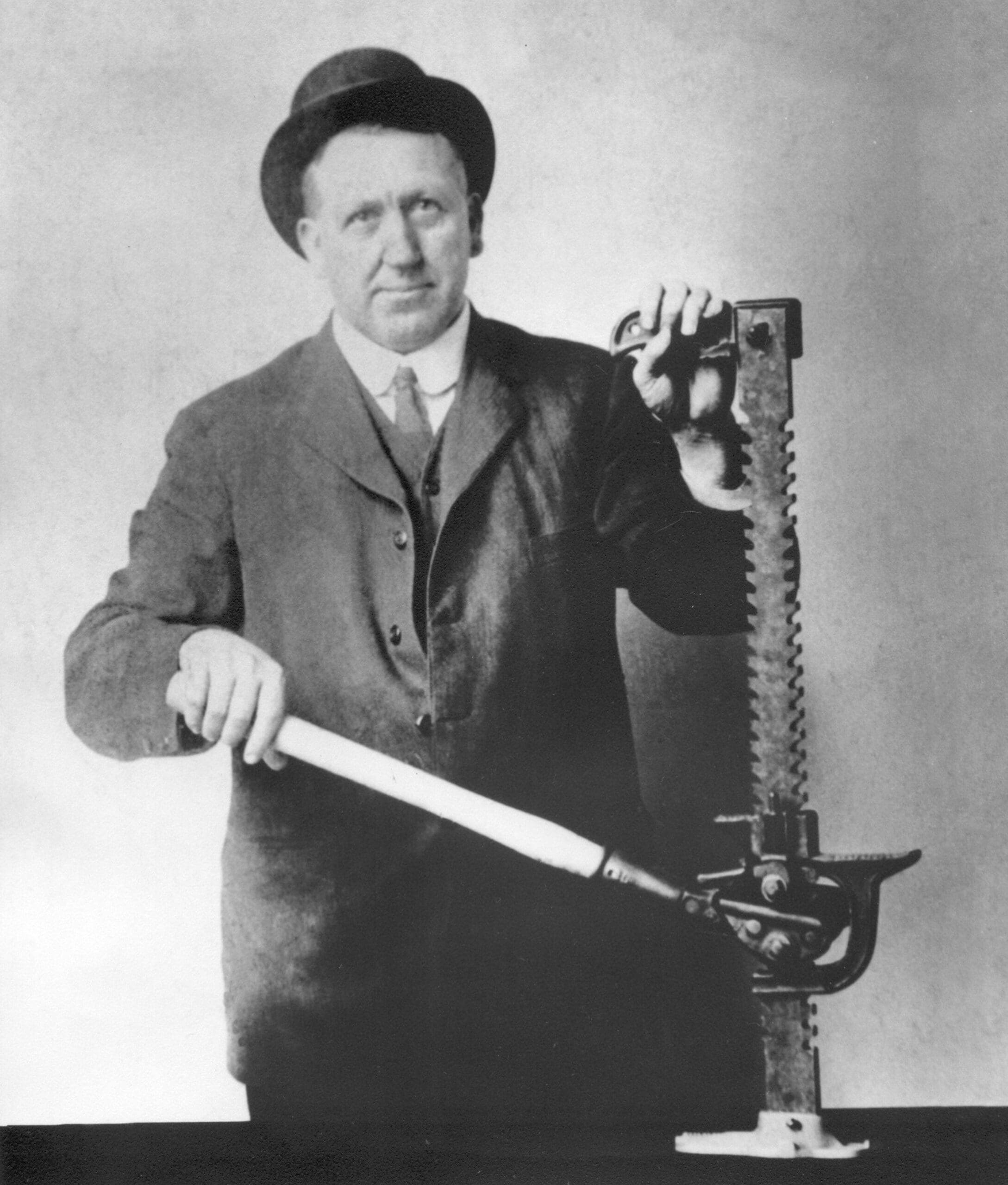 A man in a suit and hat poses with an old mechanical tool, gripping its handle. The tool stands upright on a table, featuring gears and a long metal rod with teeth, suggesting it may be an early version of a hi-lift jack.