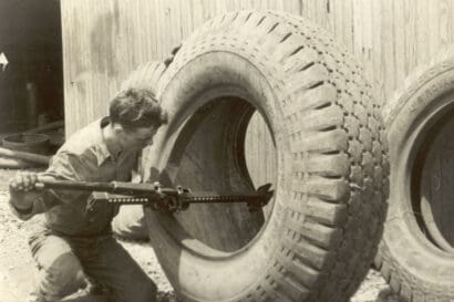 A man kneels beside a large tire, using a tool and a hi-lift to work on it. There are other large tires and a wooden building wall in the background.