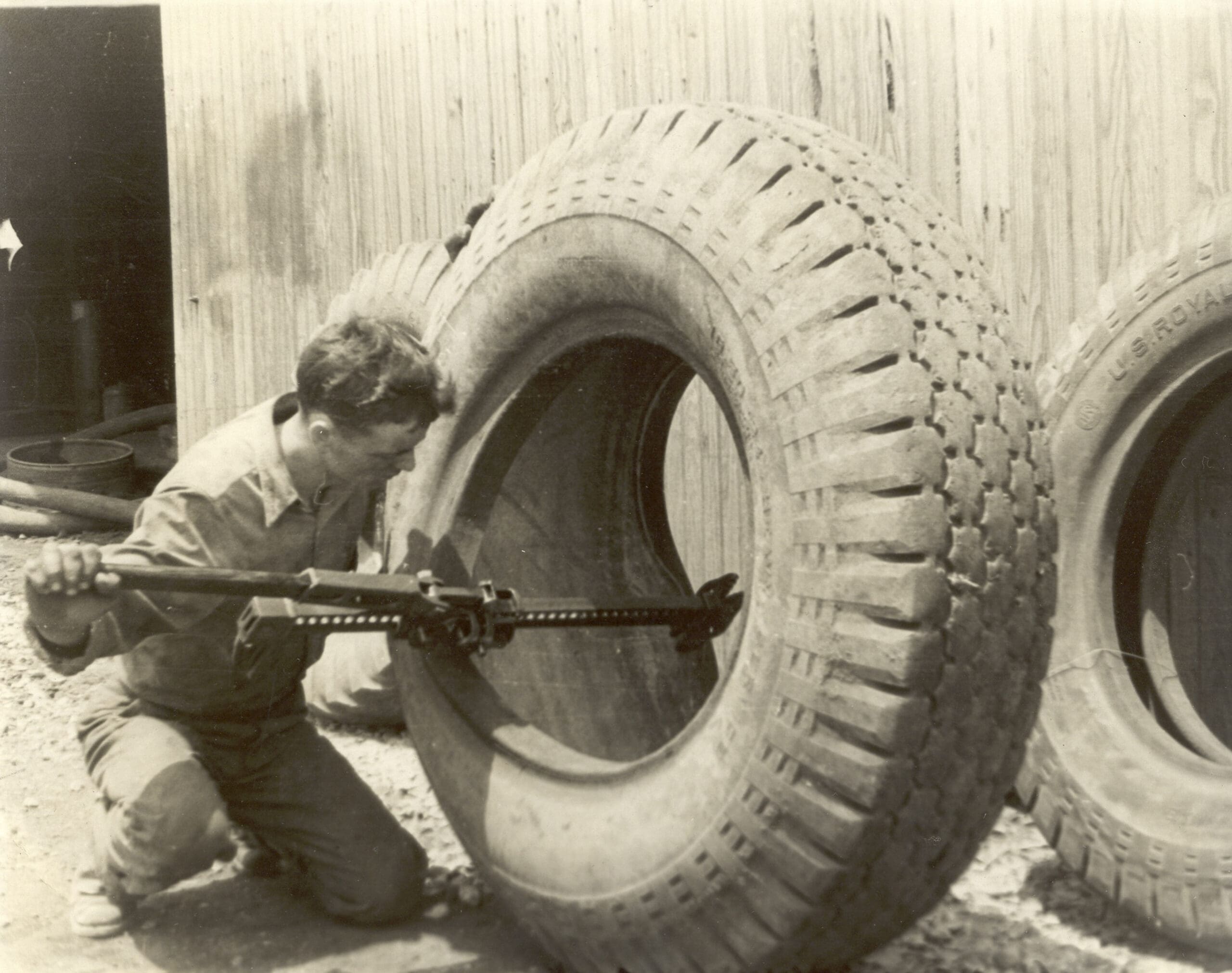 A man kneels beside a large tire, using a tool and a hi-lift to work on it. There are other large tires and a wooden building wall in the background.