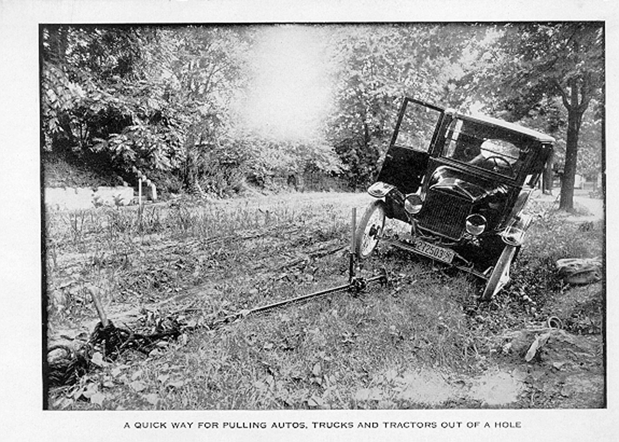 A vintage car is stuck in a ditch with its door open, being pulled out using a hi-lift jack and a rope or cable attached to a tree. Trees and grass are visible around.