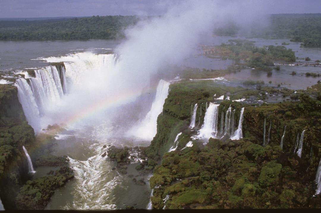 Aerial view of Iguazu Falls with mist rising and a rainbow arching over the waterfalls; lush green forest surrounds the powerful cascades—a breathtaking sight for those journeying 6,000 miles by Land Rovers to witness nature’s grandeur.