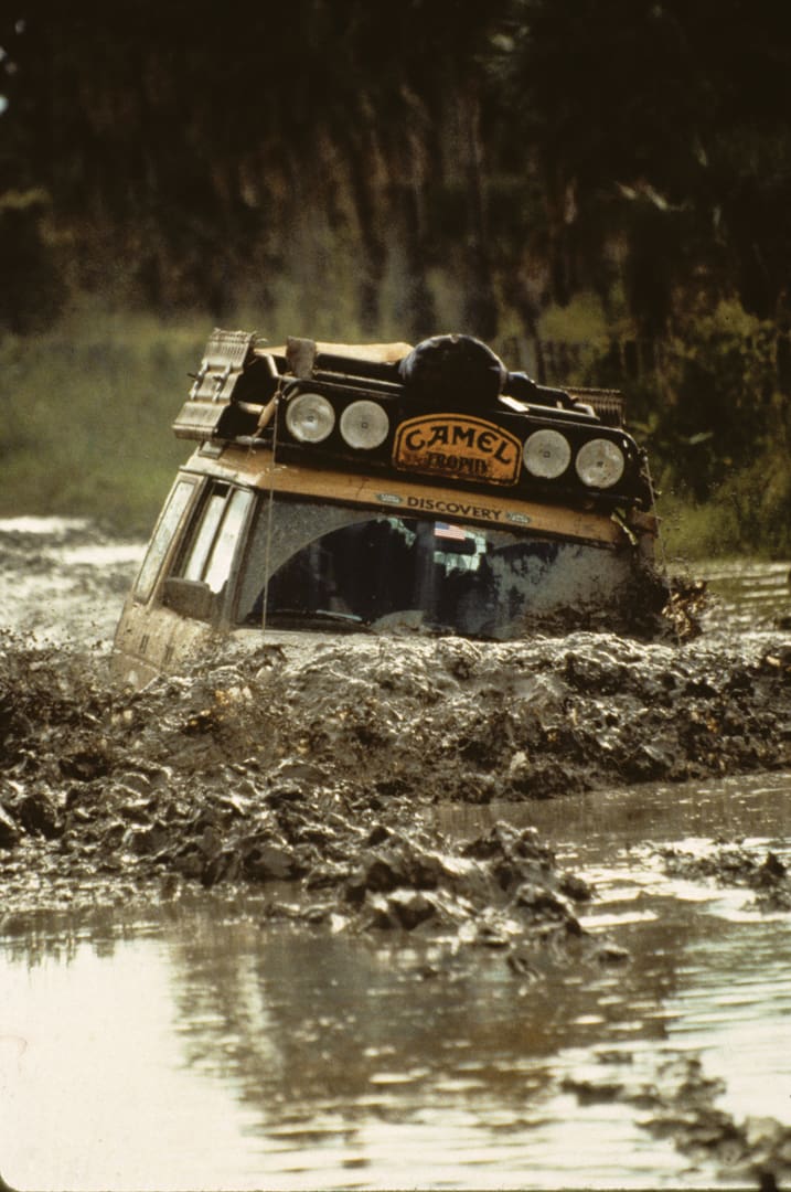 A yellow off-road Land Rover with Camel Trophy branding drives through deep, muddy water, splashing mud around as it navigates a rugged, forested terrain—just one moment in a grueling 000 Miles journey over 28 Days.