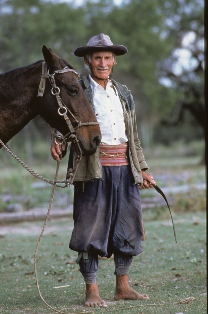 A man wearing a hat, traditional clothing, and no shoes stands next to a brown horse outdoors—holding its reins and a leather whip—set against green trees in the background. After traveling 6,000 miles in 28 days with Land Rovers, he now takes a quiet moment beside his trusted horse.