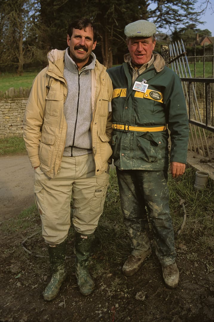 Two men in outdoor clothing and muddy boots stand side by side, smiling at the camera in a rural setting with a fence and trees in the background, their expressions reflecting the spirit of adventure that comes from a day spent exploring off the beaten path.