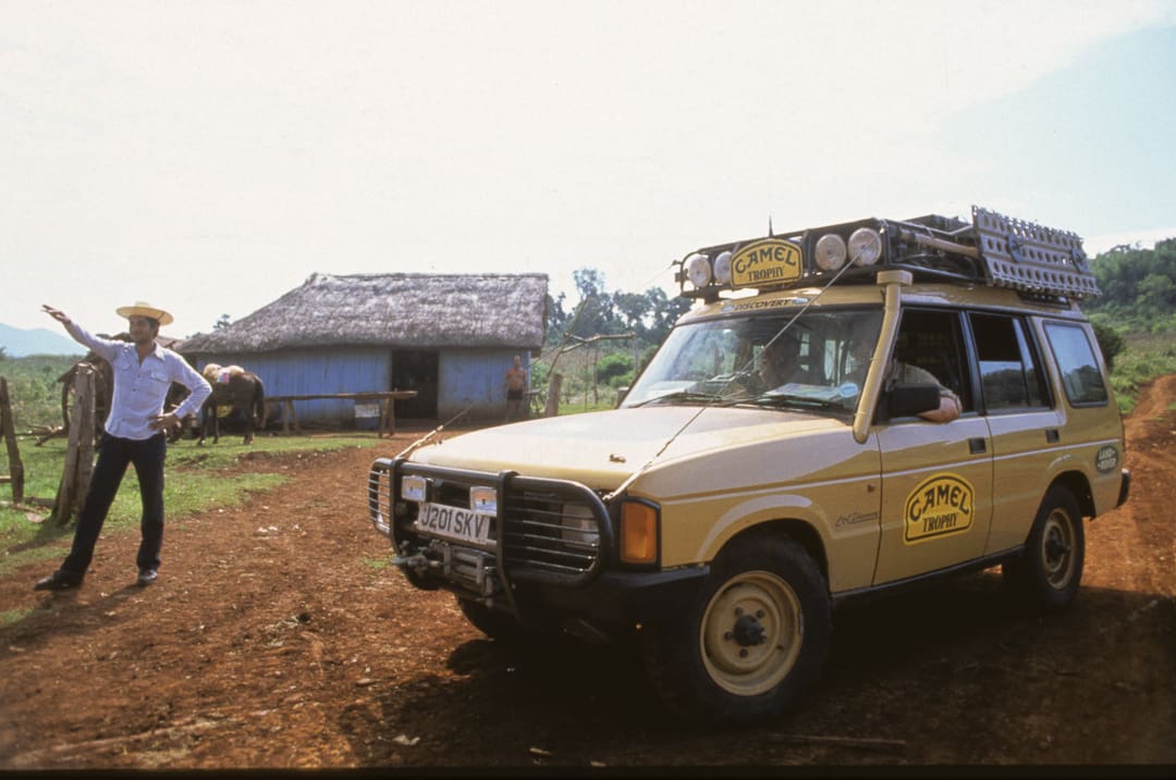 A yellow Camel Trophy Land Rover is parked on a dirt road near a thatched house, while a person wearing a cowboy hat points to the left—capturing the spirit of 6,000 miles of adventure in just 28 days.