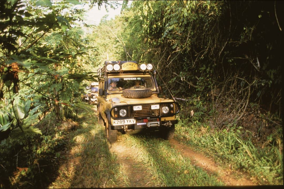 A yellow Land Rover Defender drives on a narrow, muddy jungle trail surrounded by dense green foliage, with another vehicle following behind—just one part of an epic 28 Days, 6 Land Rovers, 000 Miles adventure through untamed wilderness.