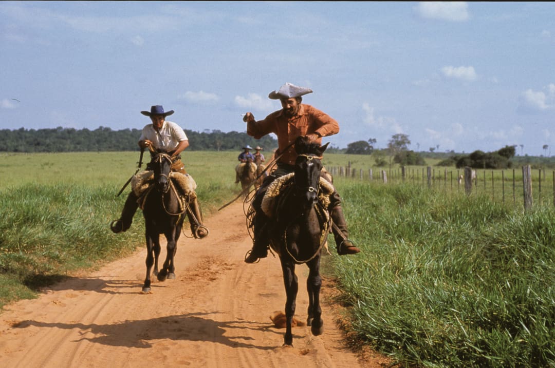 Two people ride horses on a dirt road through grassy fields, with others following in the background. Both riders wear wide-brimmed hats and rural attire, under a partly cloudy sky, as if embarking on an adventure spanning 28 days and 6,000 miles—echoes of Land Rovers exploring the wild alongside them.