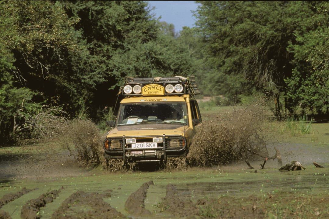 A yellow Land Rover off-road vehicle drives through a muddy, water-filled path in a forest, splashing mud and water around—ready to conquer 6,000 Miles in 28 days of adventure.