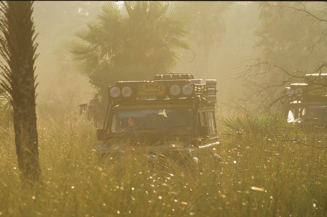 Two off-road vehicles, both classic Land Rovers, drive through tall grass and haze, surrounded by palm trees. The front vehicle has multiple lights and a yellow Camel Trophy sign on the roof. Evoking 28 days of rugged adventure and exploration across 6,000 miles, the scene appears truly adventurous and wild.