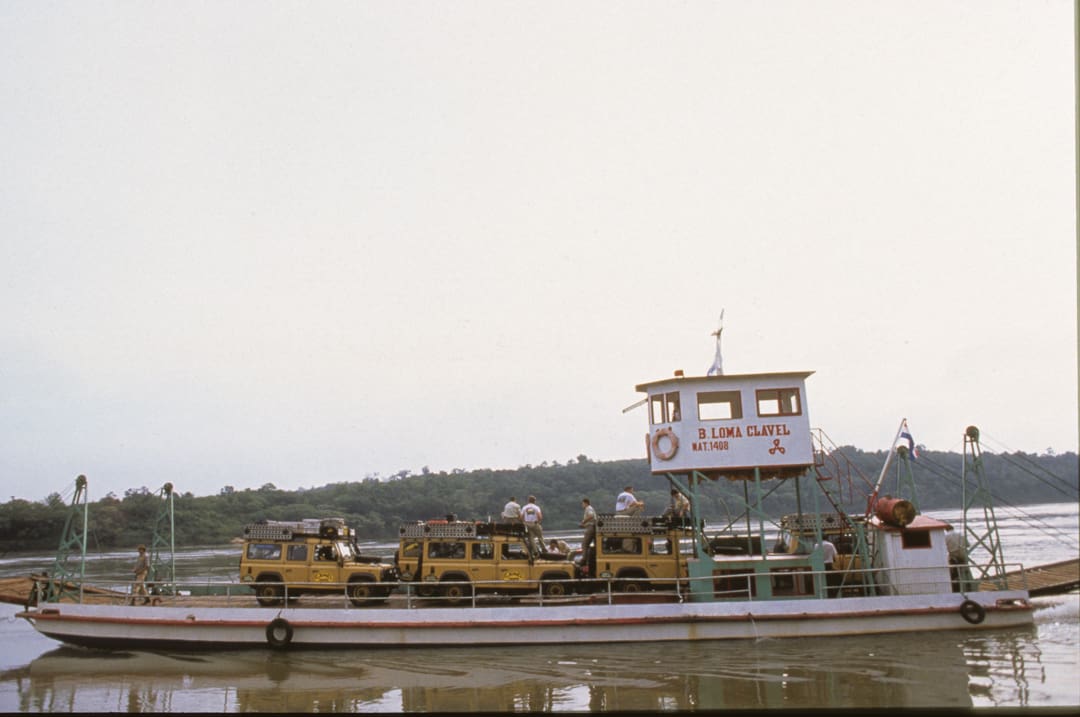 A ferry named S. LOMA CLAVEL carries several yellow Land Rovers and passengers across a wide river, with a forested shoreline in the background under a cloudy sky—a journey that could span 6,000 miles in just 28 days.