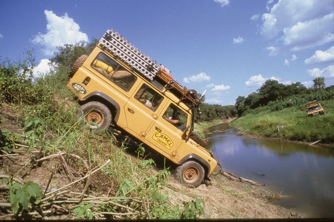 A yellow Land Rover with roof gear drives down a steep, grassy riverbank toward water. Another similar vehicle is visible in the background under a blue sky with scattered clouds—two explorers on their 6,000 miles journey over 28 days across rugged terrain.