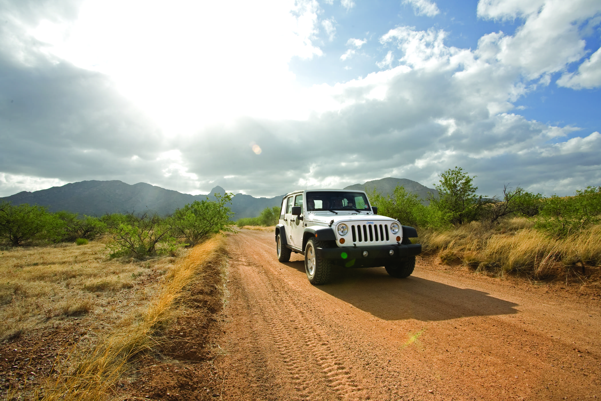 A white Jeep drives on a dirt road through Arizona’s dry, grassy borderlands, with bushes and mountains in the background under a partly cloudy sky and bright sunlight.