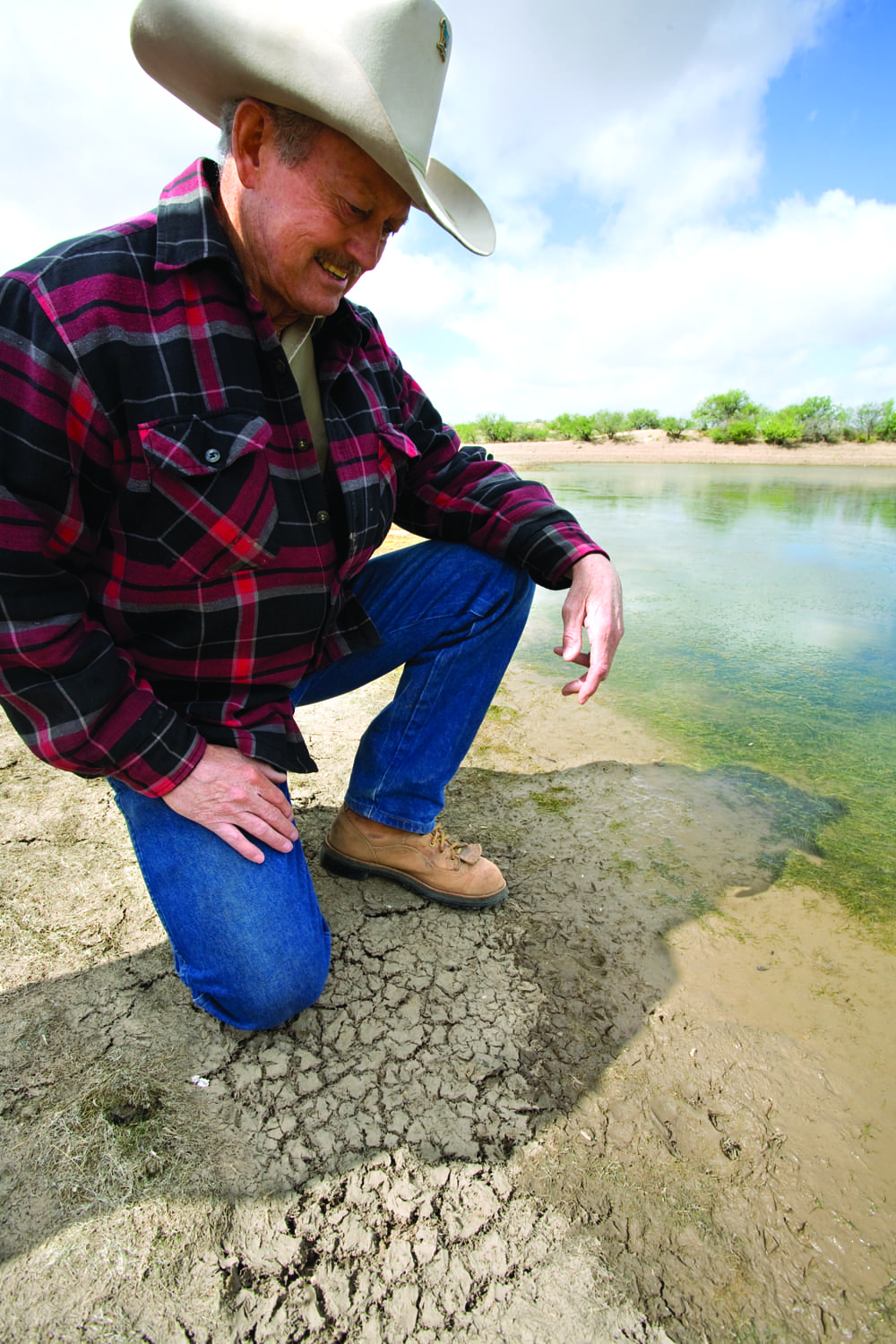 A person in a cowboy hat and plaid jacket kneels on dry, cracked ground near a body of water in the Arizona borderlands, looking down and smiling, with greenery in the background.