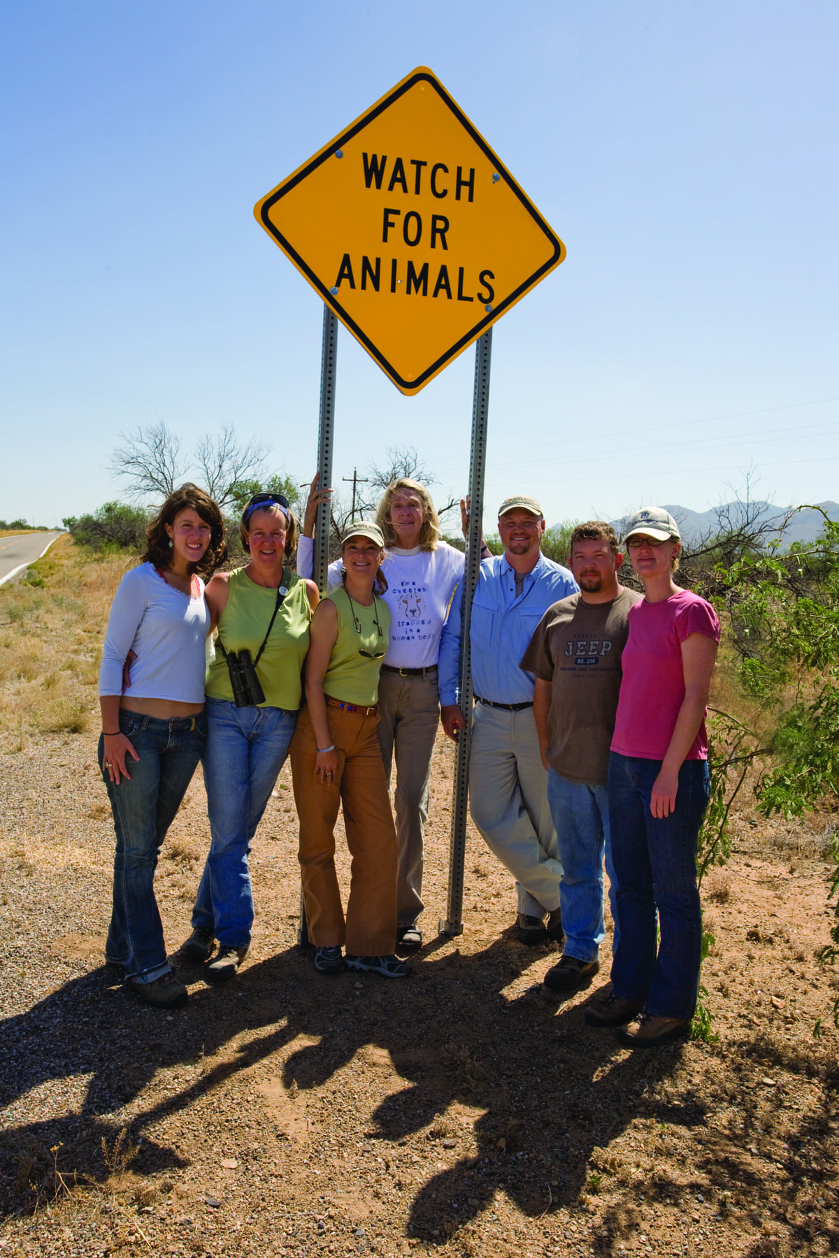 Seven people stand together on a roadside under a yellow diamond sign that reads Watch for Animals in the sunny Arizona borderlands, desert vegetation behind them—an area known for elusive wild cats like the Jaguar.