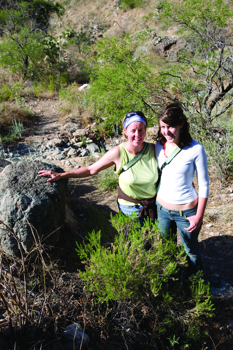 Two women stand on a rocky, sunlit hiking trail in the Arizona borderlands, smiling at the camera. One has her arm around the other and touches a large rock, surrounded by green shrubs and trees where wild cats may roam.