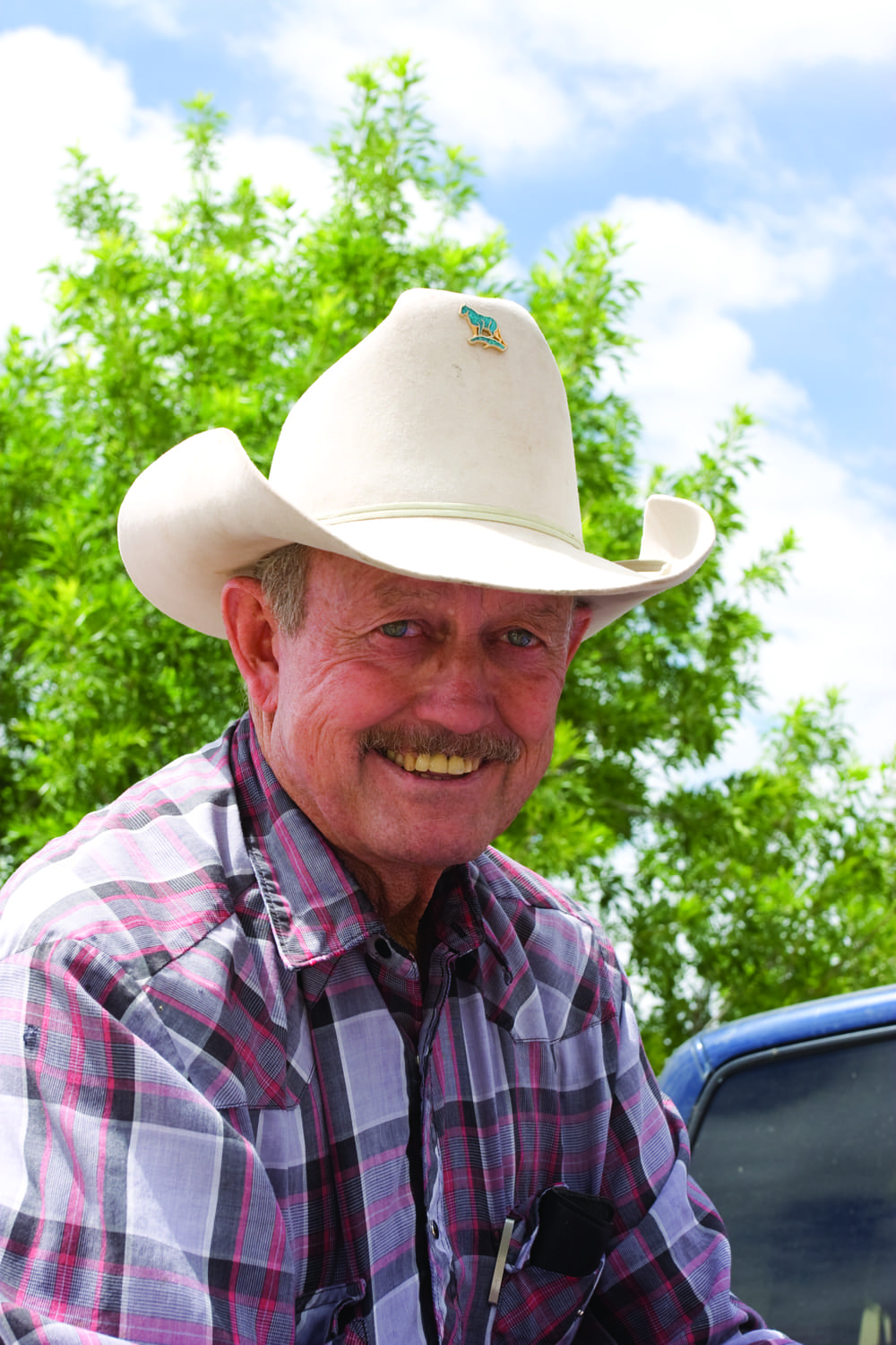 Smiling older man in a white cowboy hat and plaid shirt poses outdoors in the Arizona borderlands, with green trees and a blue sky behind him.