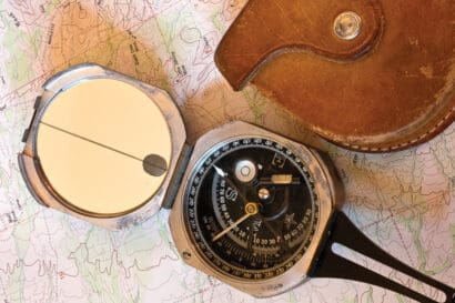 A metal Brunton Pocket Transit compass with a mirror and sighting line rests on a topographic map next to a brown leather case.