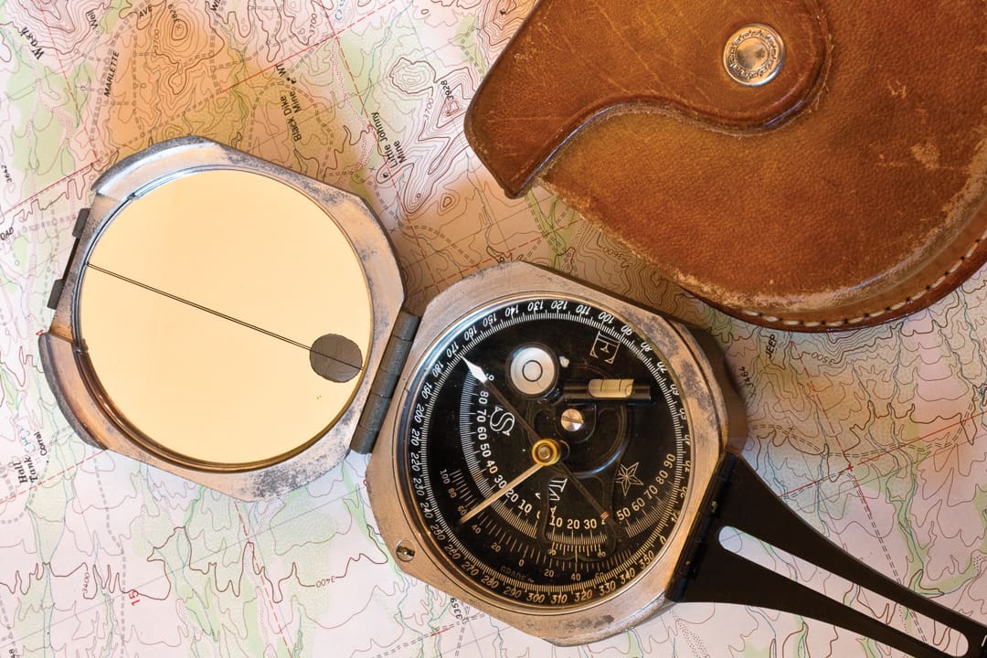 A metal Brunton Pocket Transit compass with a mirror and sighting line rests on a topographic map next to a brown leather case.