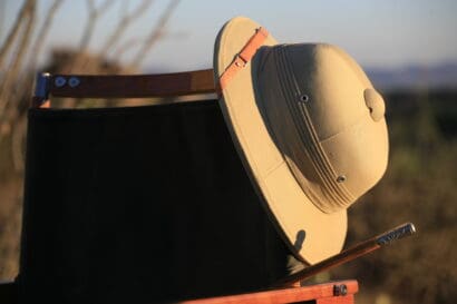 A tan pith helmet—a true classic kit essential—rests on the back of a wooden chair, with blurred outdoor scenery and warm sunlight in the background.