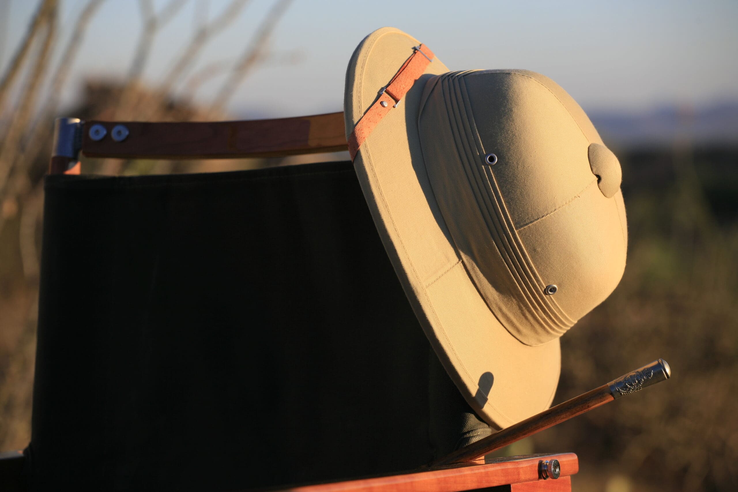 A tan pith helmet—a true classic kit essential—rests on the back of a wooden chair, with blurred outdoor scenery and warm sunlight in the background.