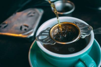 Hot water is being poured through a metal tea strainer filled with loose leaf tea into a white cup with a green saucer, creating the perfect brew for any classic tea kit.