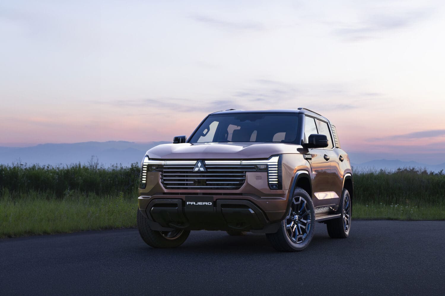 A brown Mitsubishi Pajero SUV is parked on a road at sunset, with grass and mountains in the background. The iconic Mitsubishi Pajero features modern LED headlights and a bold front grille design.