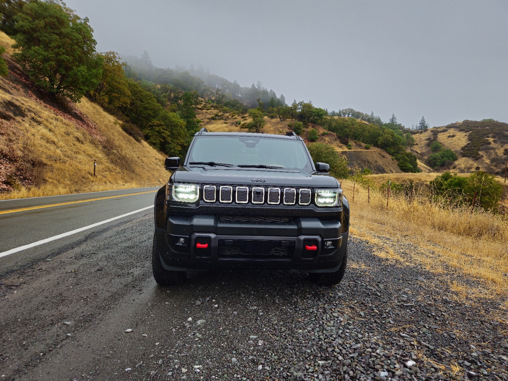 A black Jeep Recon SUV is parked on the side of a winding mountain road with dry grass, trees, and hills in the background under an overcast sky.