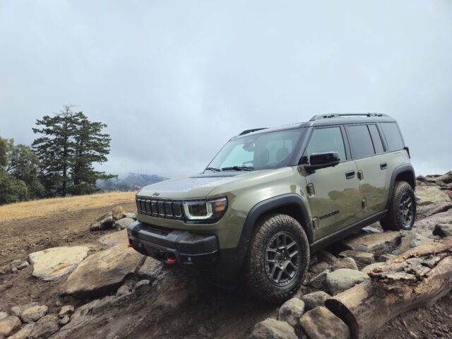 A green Jeep Recon SUV with rugged tires is parked on uneven rocky terrain outdoors, with cloudy skies and trees in the background.