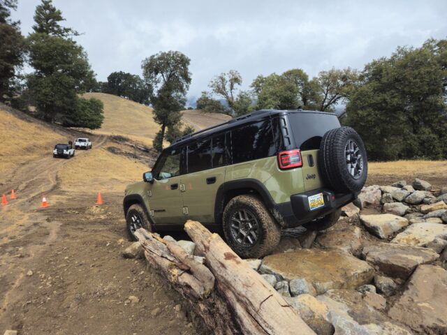 A green Jeep Recon SUV drives over rocks and wooden logs on a dirt trail, with orange cones and another vehicle in the background, surrounded by trees and grassy hills.