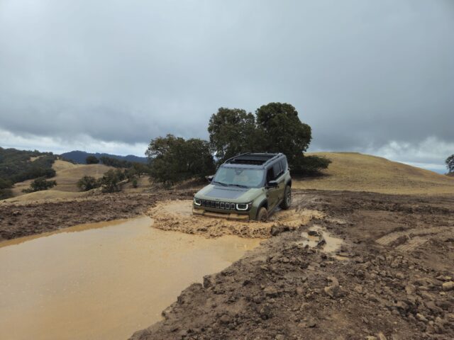 A green Jeep Recon drives through a large muddy puddle on a rural dirt road, surrounded by hills and scattered trees under a cloudy sky.