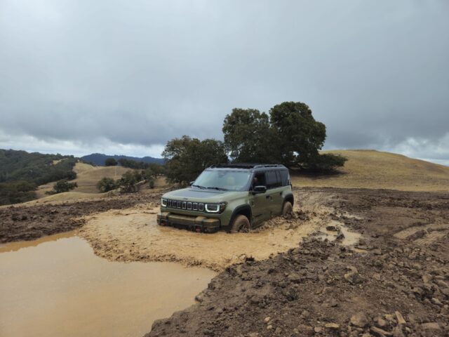 A green Jeep Recon off-road vehicle is stuck in a large, muddy puddle on a dirt trail. Rolling hills, scattered trees, and a cloudy sky are visible in the background.