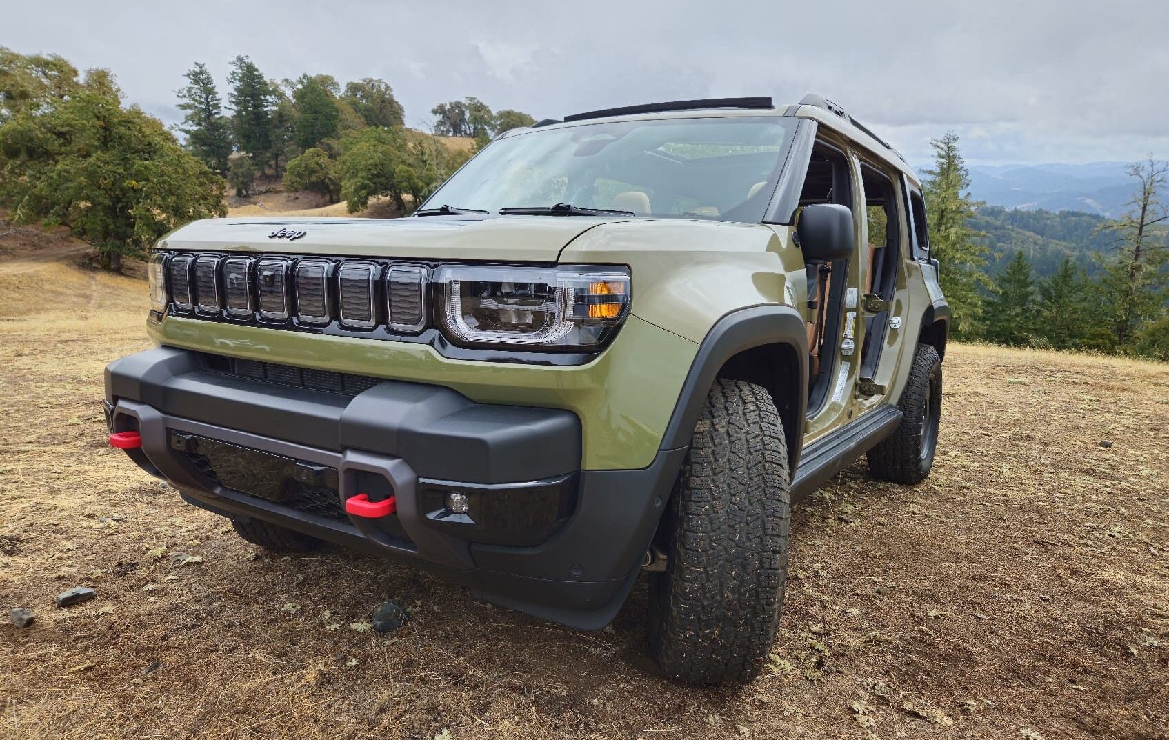 A green Jeep Recon SUV with its doors removed is parked on a dry, grassy hill with trees and mountains in the background under a cloudy sky.